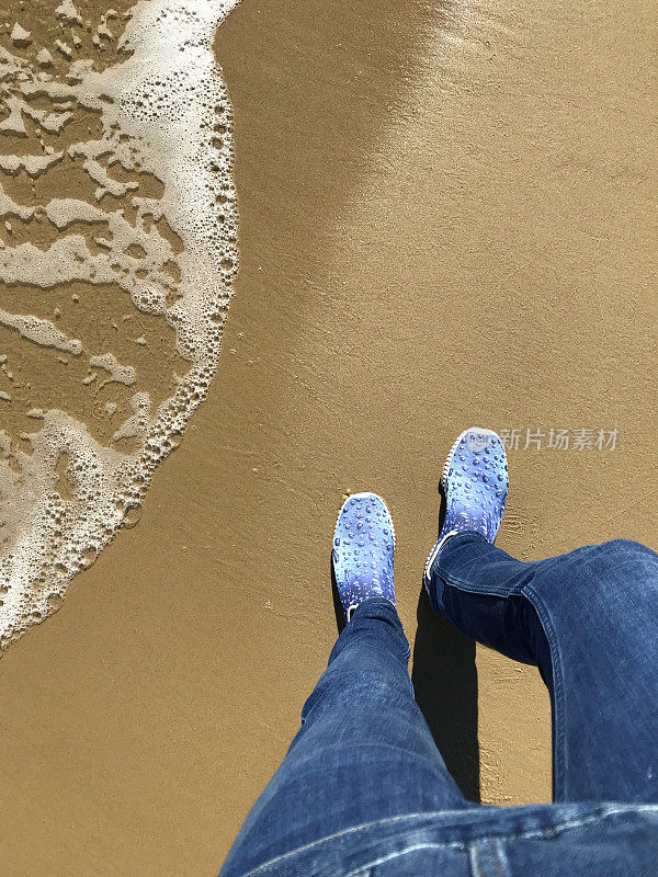 白色的英国人的脚穿着蓝色的海滩鞋和紧身牛仔裤在夏天度假，金色的沙子被海浪拍打着海岸线，步行池水鞋海滨脚印站在海滩海岸，泡沫/泡沫来自潮水